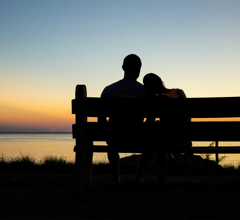 Showing a man and a woman seated on a bench looking out at the sunset over the water.